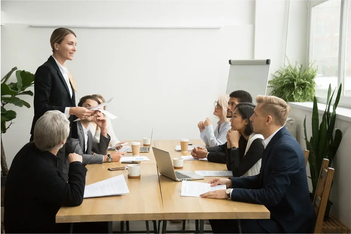 Una reunión de negocios en una oficina luminosa, con un presentador de pie y colegas sentados alrededor de una mesa, interactuando y tomando notas.